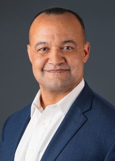 Headshot of a man in a blue suit against a dark grey background.