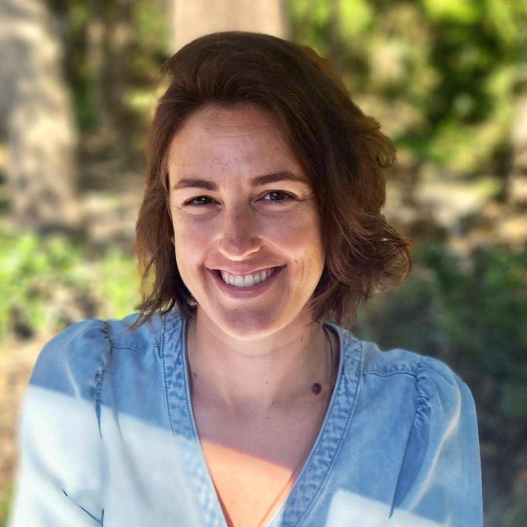 A woman smiling at the camera with a natural outdoor background, wearing a blue shirt, surrounded by trees.