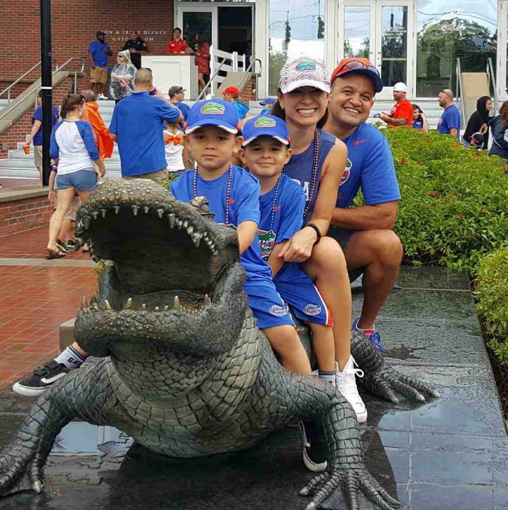 A family in matching blue shirts and Gators hats poses on a bronze alligator statue at the University of Florida.