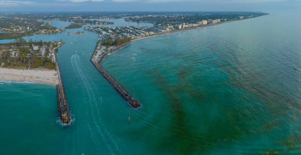 Aerial view of Jupiter Inlet, Florida, showing the inlet leading into a waterway with boats, surrounded by residential areas and greenery on both sides. Red tide is present in the water.