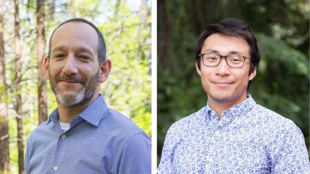 Two individuals posing outdoors, one wearing a blue shirt and the other in a patterned blue shirt, both smiling in a natural setting.