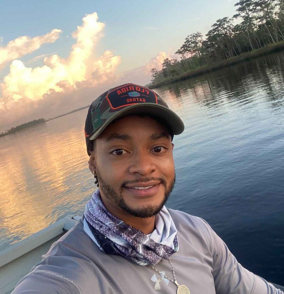 A man in a camo hat smiles on a boat. Calm water reflects the sky with fluffy clouds and distant trees.