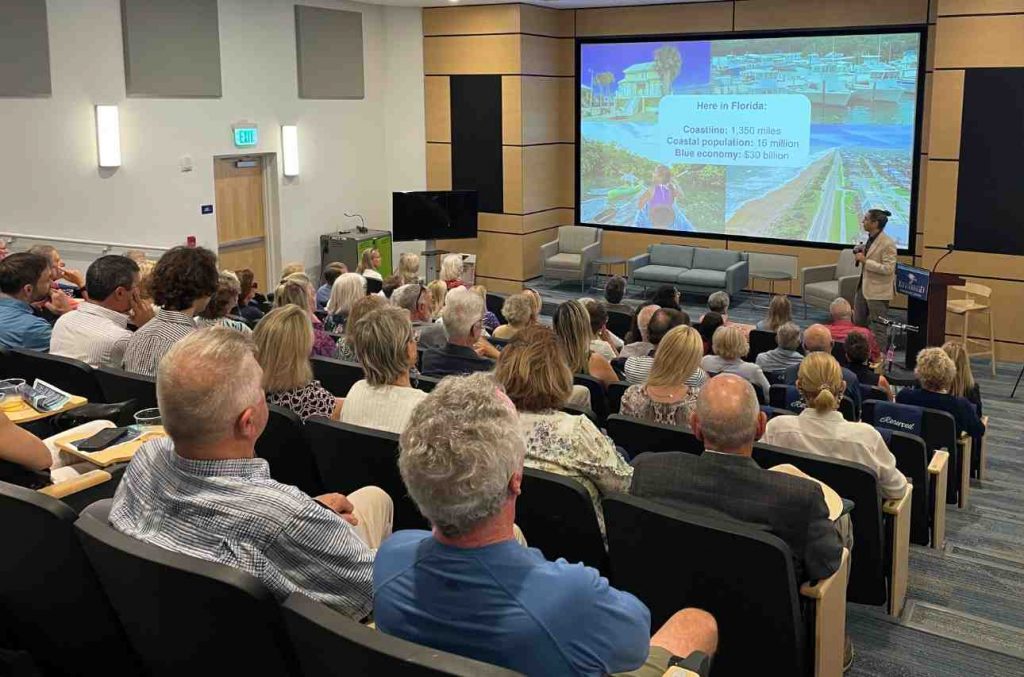 A speaker presents to a seated audience in a modern lecture hall. The slide shows Florida's coastline data. The room is filled with engaged attendees.