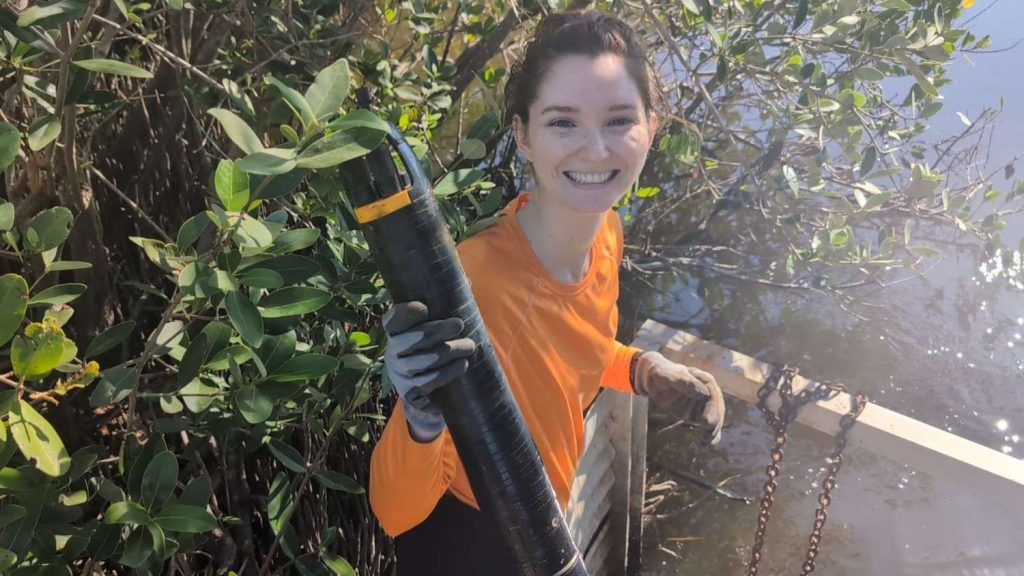 A person wearing an orange top and gloves is smiling while standing near a body of water, holding a cylindrical object. Dense green foliage and a partially submerged structure with chains are in the background.