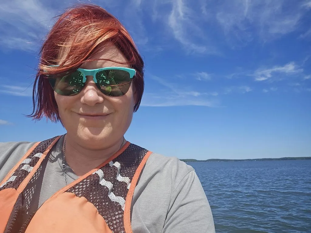 Nina Stark smiles for a selfie while wearing sunglasses and an orange life vest, with open water and a blue sky behind her.